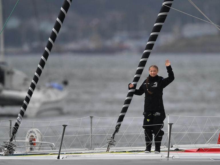 Greta Thunberg steht auf dem Deck des Segelbootes und winkt Richtung Kamera.