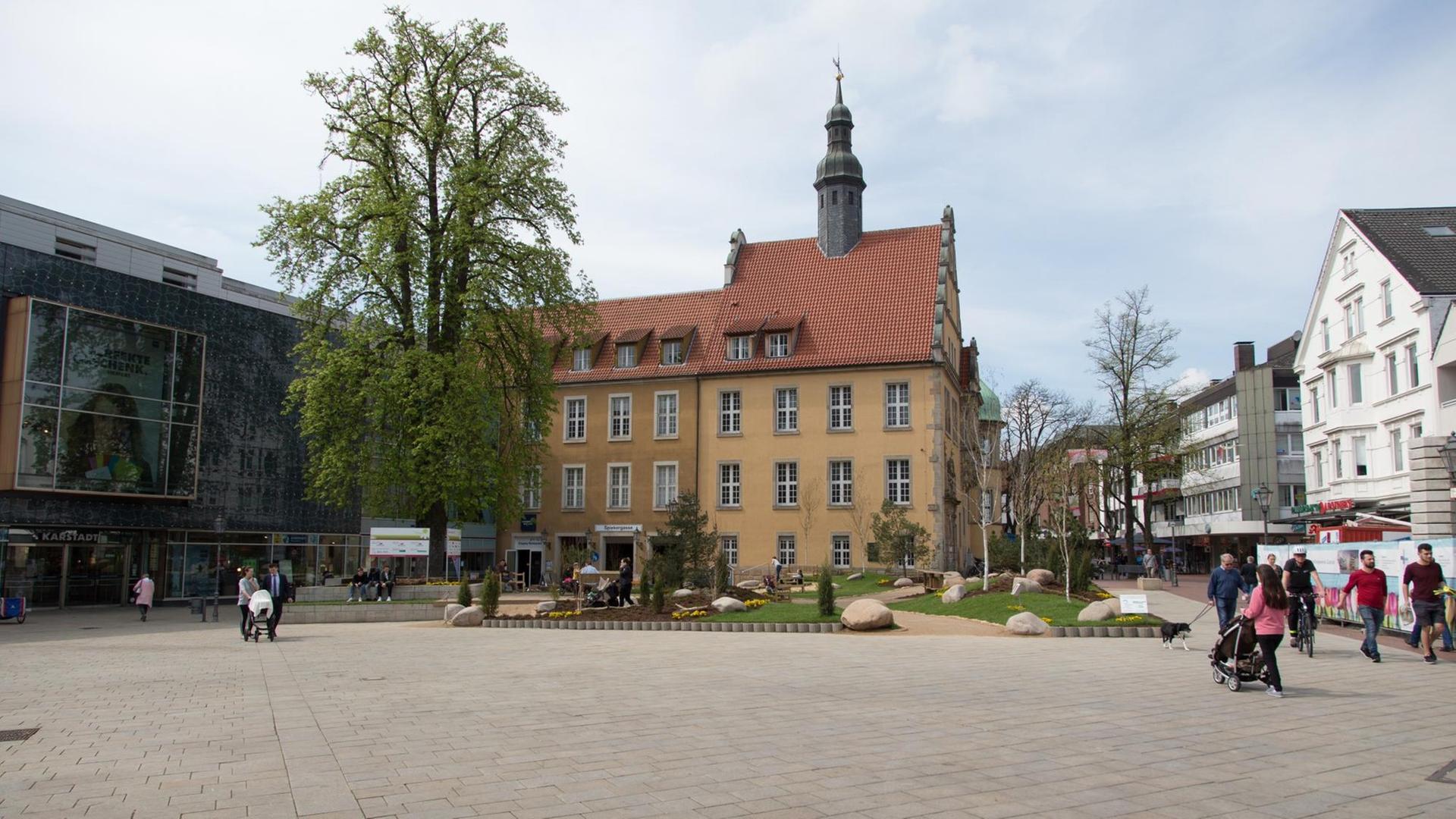 Blick auf einen städtischen Platz, im Hintergrund ein historisches Gebäude und ein Baum sowie einzelne Passanten