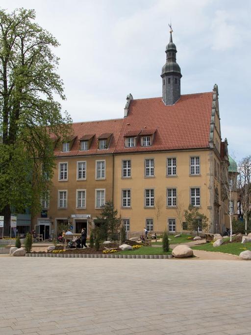 Blick auf einen städtischen Platz, im Hintergrund ein historisches Gebäude und ein Baum sowie einzelne Passanten