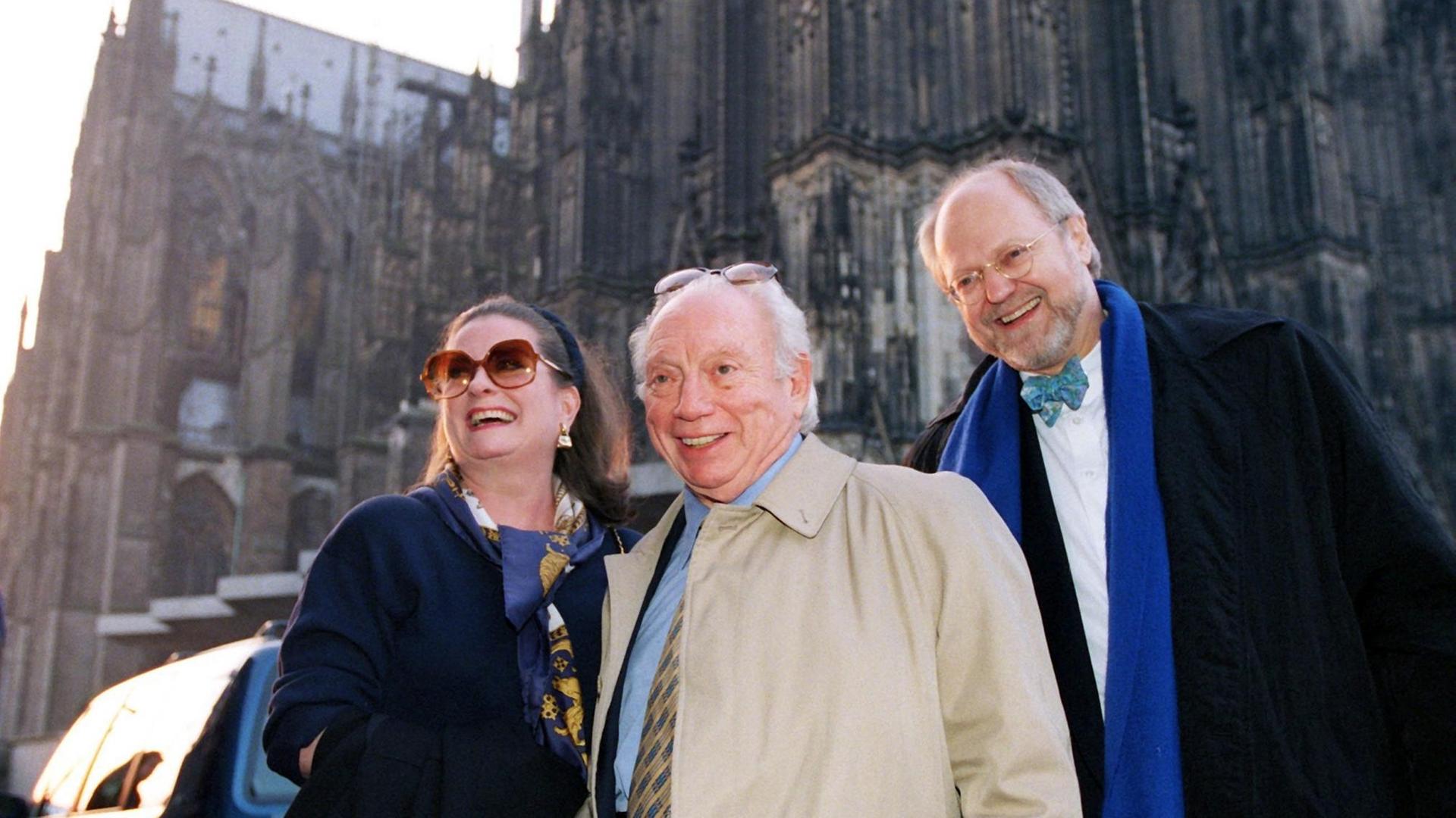 Ein grauhaariger alter Mann mit Brille auf der Stirn steht, umgeben von zwei anderen, wie er lachenden, Menschen, vor dem Kölner Dom.