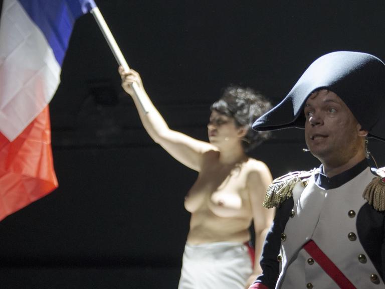 Ein Darsteller im Napoleonskostüm steht auf der Bühne. Im Hintergrund sieht man eine barbusige Frau, die die Frankreichflagge schwenkt.