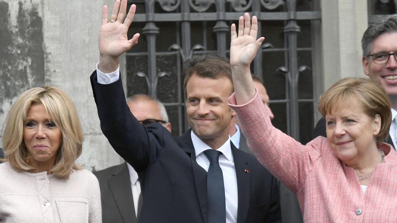 Macron steht zwischen seiner Frau und Bundes-Kanzlerin Merkel. Sie winken den Zuschauern zu.
