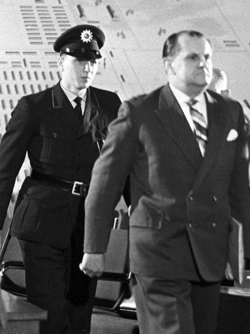 Der Angeklagte Oswald Kaduk wird von drei Polizisten in einen Gerichtssaal geführt. Der Angeklagte Oswald Kaduk wird von drei Polizisten in einen Gerichtssaal geführt.