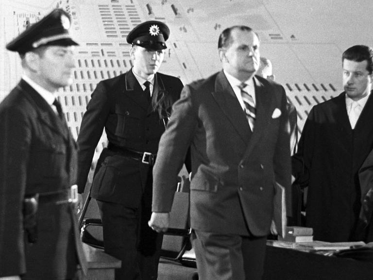 Der Angeklagte Oswald Kaduk wird von drei Polizisten in einen Gerichtssaal geführt. Der Angeklagte Oswald Kaduk wird von drei Polizisten in einen Gerichtssaal geführt.