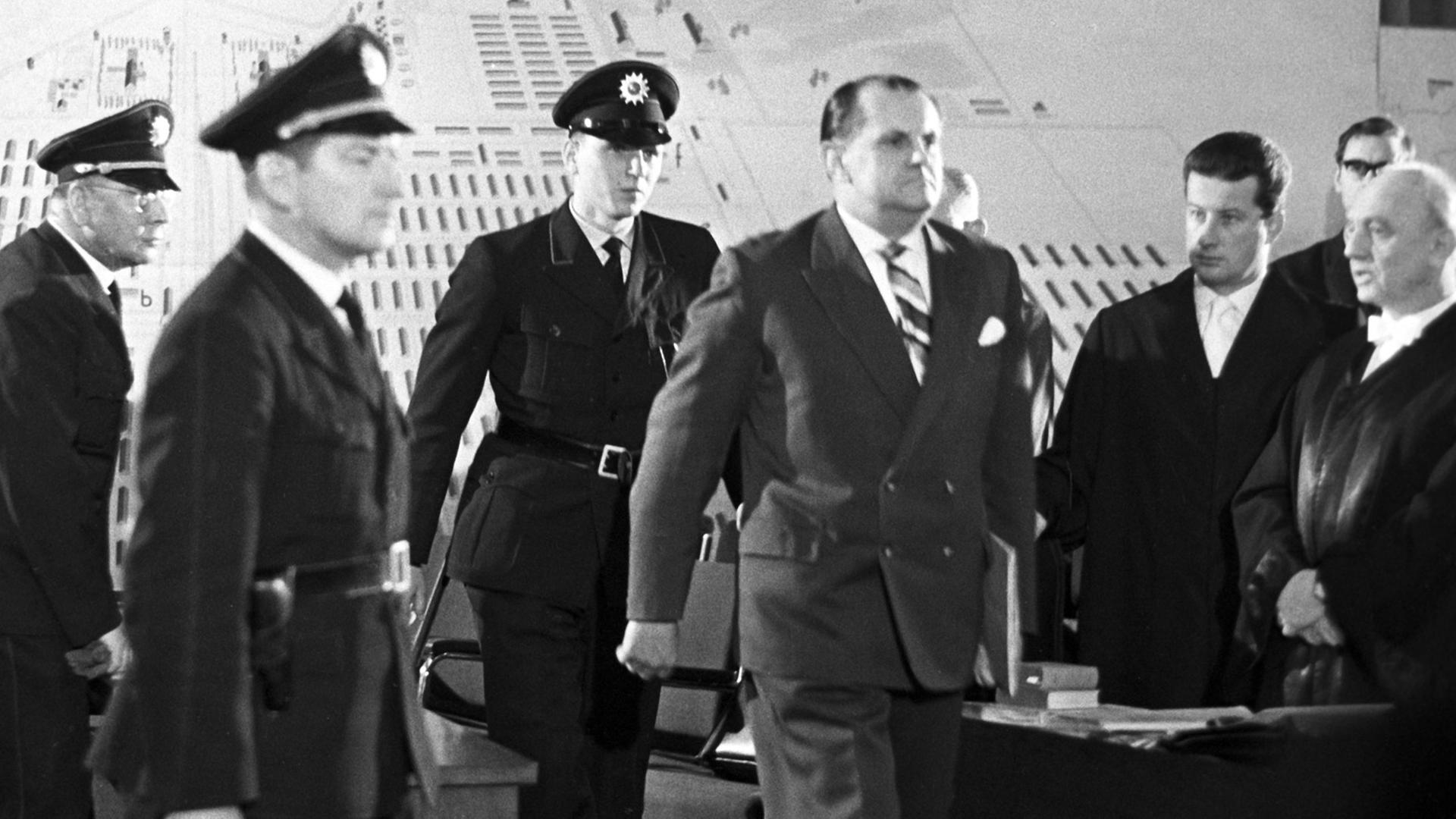 Der Angeklagte Oswald Kaduk wird von drei Polizisten in einen Gerichtssaal geführt.