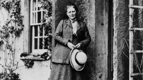 Historische Schwarz-Weiß Aufnahme einer Frau in mittleren Jahren. Sie steht bekleidet mit Rock und Anzugjacke, einen Hut in der Hand, vor einem englischen Landhaus. Es ist die Kinderbuchautorin Beatrix Potter im Jahr 1905.