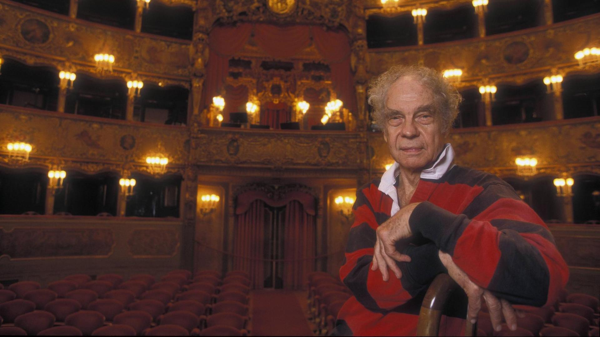 Der Choreograf Merce Cunningham sitzt im La Fenice Theater in Venedig auf einem Stuhl, im Hintergrund ist der Theatersaal mit rot bezogenen Stühlen und goldfarbenem Stuck zu sehen.