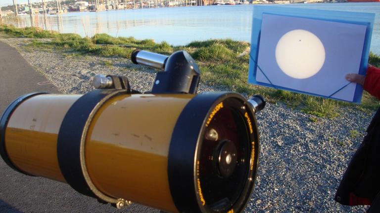 Auf einem Feldweg steht ein Hobbyteleskop. Das Okular  projiziert auf ein Blatt Papier. Vor der Sonne ist die Venus als winziger schwarzer Punkt sichtbar.
