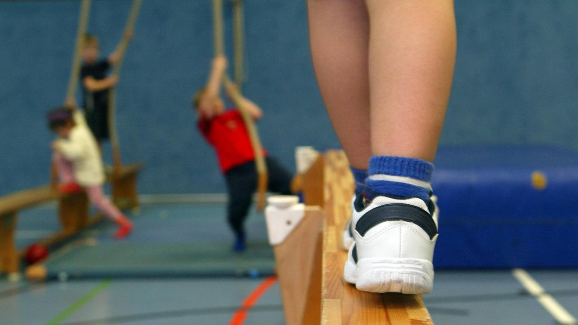 Ein Kind balanciert auf einer Bank, im Hintergrund hangeln andere Kinder während des Kinderturnens an Seilen.