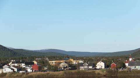 Eine kleine Siedlung in der Finnmark, ganz im Norden Norwegens. Bunte Holzhäuser stehen in einer bewaldeten Hügellandschaft und spiegeln sich in einem Fluss.