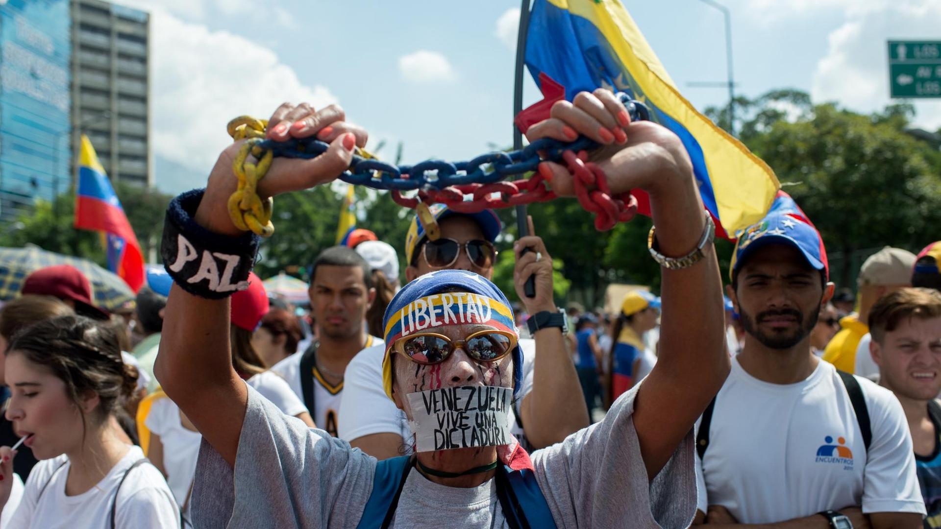 Ein Gegner von Präsident Maduro steht vor weiteren Demonstranten. Er hält eine Kette in seinen Händen; Zudem trägt er ein Stirnband mit der Aufschrift "Libertad". Das heißt "Freiheit".