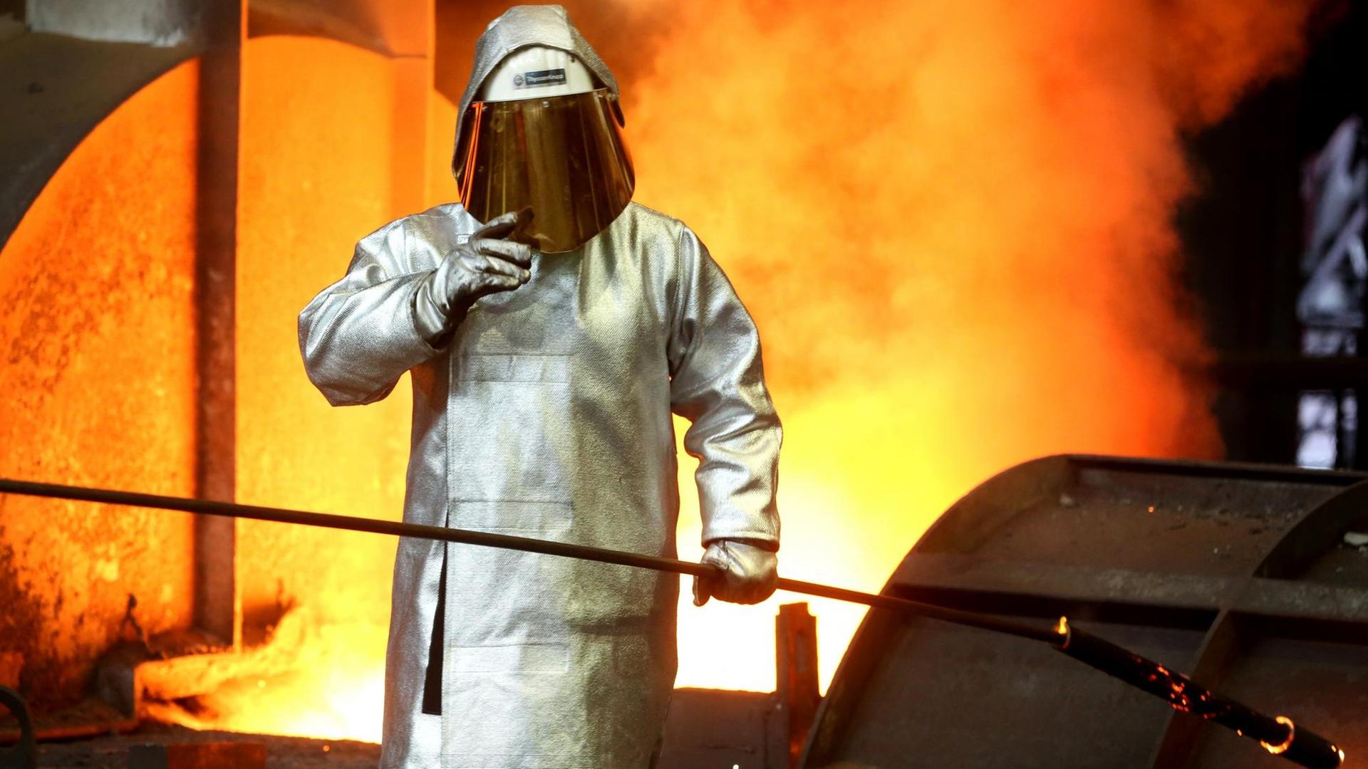 Ein Stahlarbeiter prüft am Hochofen 8 bei ThyssenKrupp in Duisburg (Nordrhein-Westfalen) die Stahlqualität nach dem Abstich. Im Hintergrund sind Glut und Flammen zu sehen.
