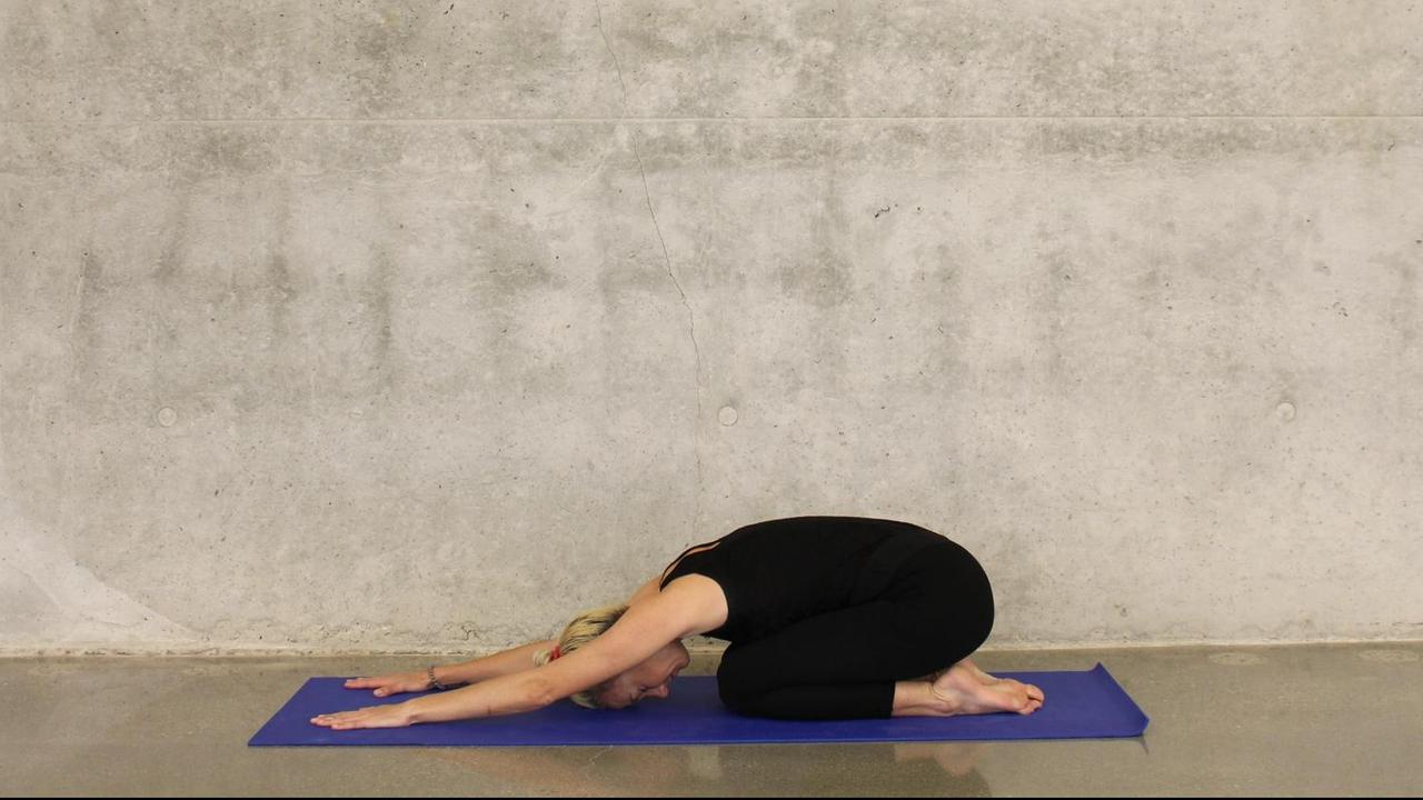 Eine sportliche Frau macht eine Yoga-Übung auf einer blauen Matte vor einer Sichtbeton-Wand.