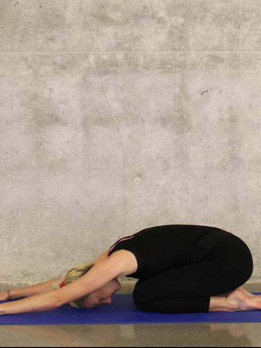 Eine sportliche Frau macht eine Yoga-Übung auf einer blauen Matte vor einer Sichtbeton-Wand.