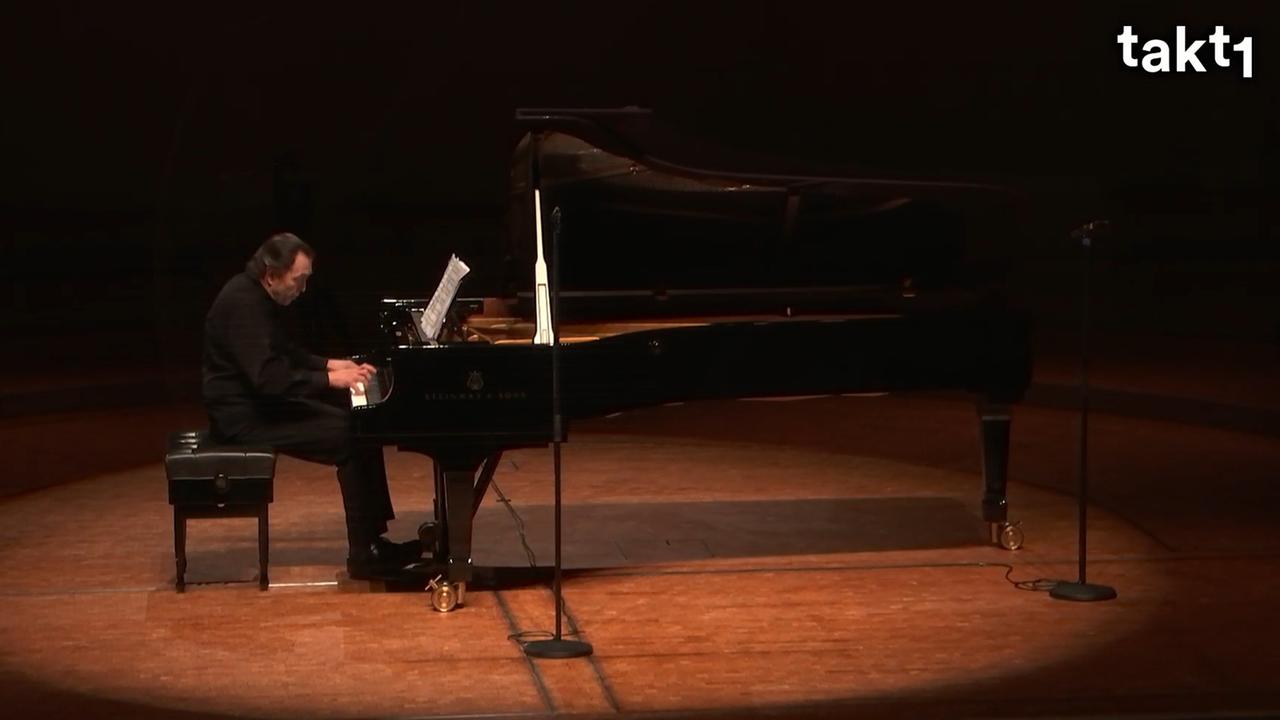 Ein Pianist sitzt an einem langen Konzertflügel. Er ist ganz schwarz gekleidet, der Flügel ist auch schwarz. Er blickt konzentriert auf die Noten vor ihm.