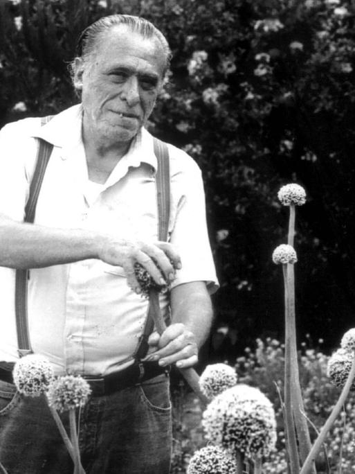 Historische schwarz-weiss Aufnahme des Schriftstellers Charles Bukowski in einem Garten, er streichelt eine phallisch aussehende Pflanze.