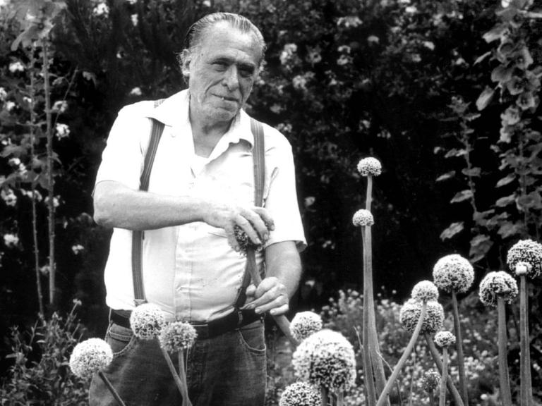 Historische schwarz-weiss Aufnahme des Schriftstellers Charles Bukowski in einem Garten, er streichelt eine phallisch aussehende Pflanze.