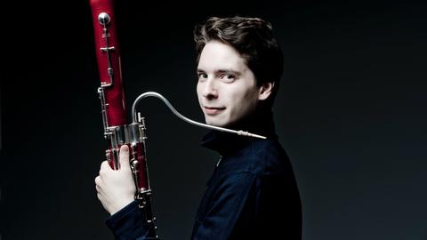 Ein dunkel gekleideter, junger Mann mit braunen Haaren blickt seitlich in die Kamera, mit Fagott in seiner linken Hand.