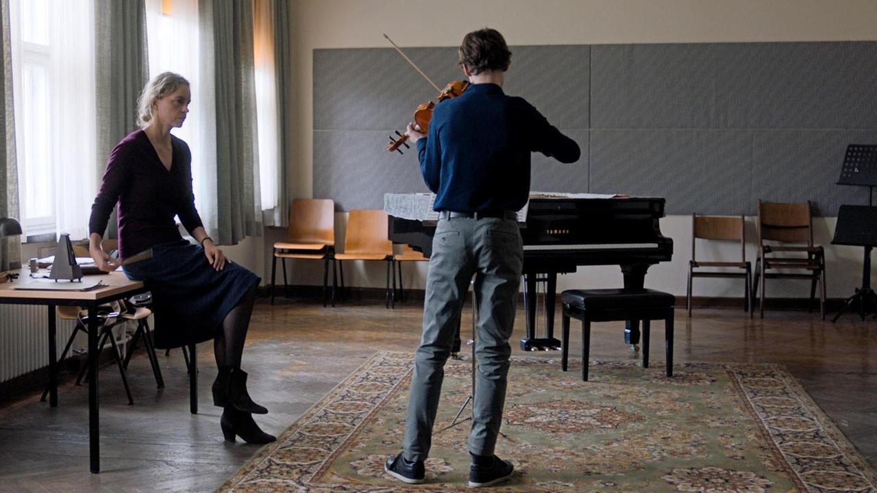 Eine Frau schaut streng auf einen Mann, der in der Mitte eines großen Raumes steht und Geige spielt.
