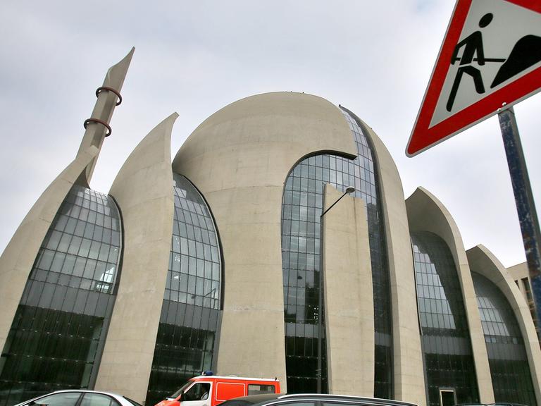 Ansicht der Kölner Zentralmoschee, eines geschwungenen Gebäudes mit Kuppel aus Beton und Glas, davor Autos auf einer Straße und ein Baustellenschild