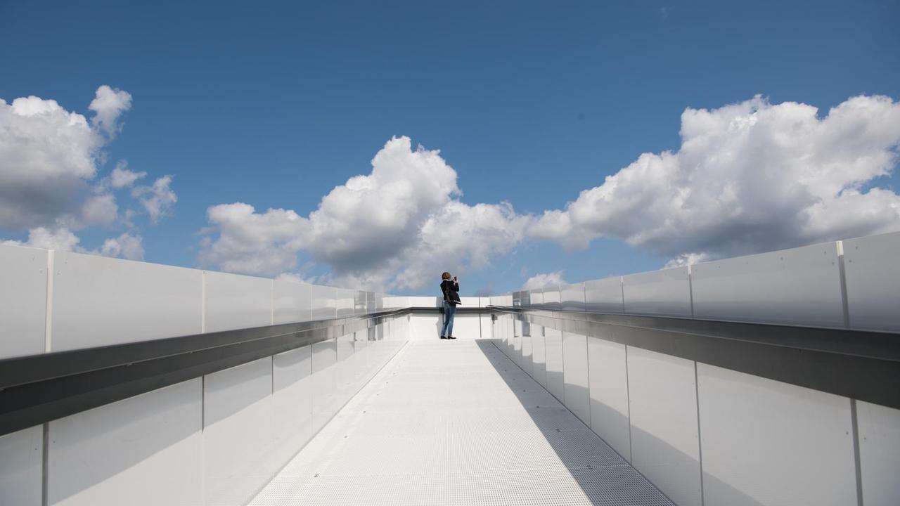 Eine Frau steht bei leicht bewölktem Wetter auf dem "Skywalk" auf dem Dach des "Futurium". 