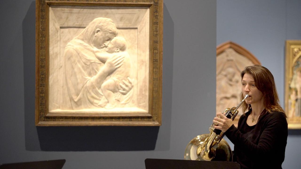 Eine Hornistin spielt inmitten eines Museums, neben ihr ein Relief, das eine Frau vertieft in die Umarmung eines Kindes zeigt.