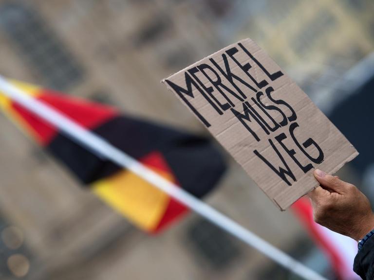 Eine Hand hält ein Schild mit der Aufschrift "Merkel muss weg" in die Höhe, dahinter eine deutsche Flagge.
