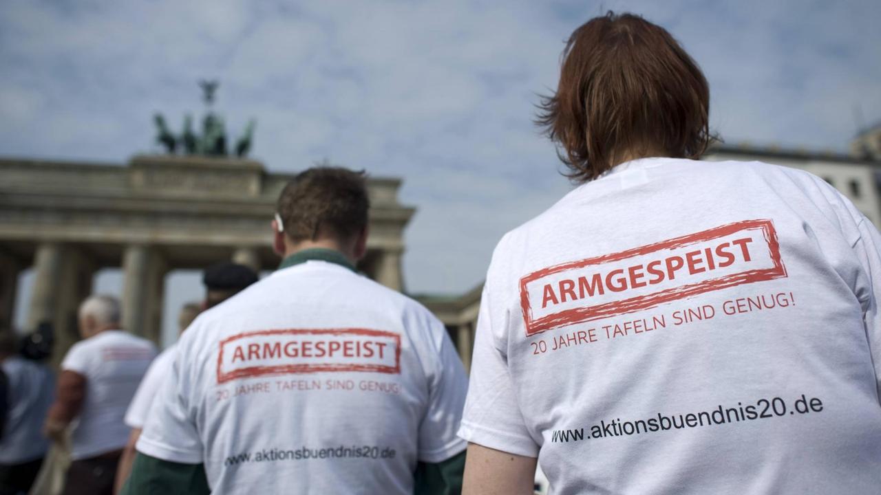 "Armgespeist! 20 Jahre Tafeln sind genug" steht auf den T-Shirts zweier Männer bei einem Protest vor dem Brandenburger Tor. Anlass ist das 20-jährige Bestehen der Berliner Tafel, der ersten in Deutschland.