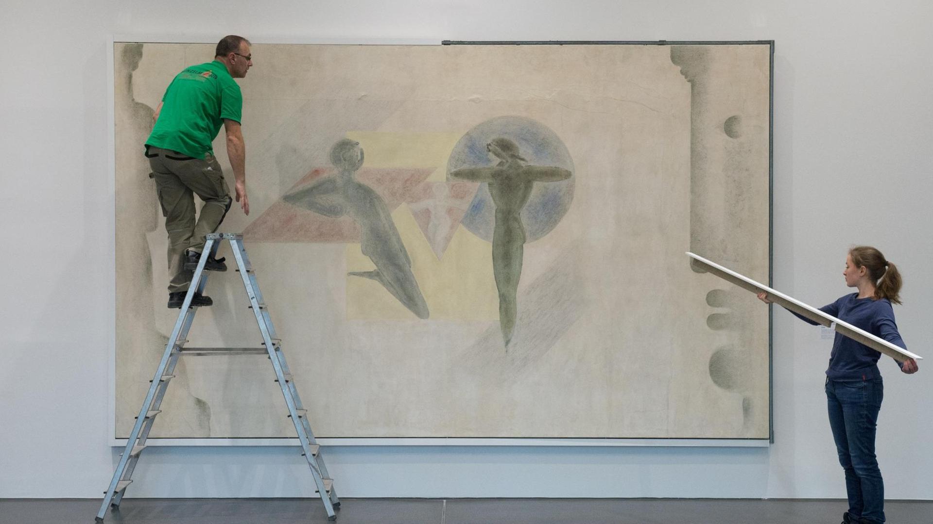 Ein Mann auf einer Leiter und eine Frau mit einem Brett in der Hand stehen vor einem Wandgemälde in einer Galerie.
