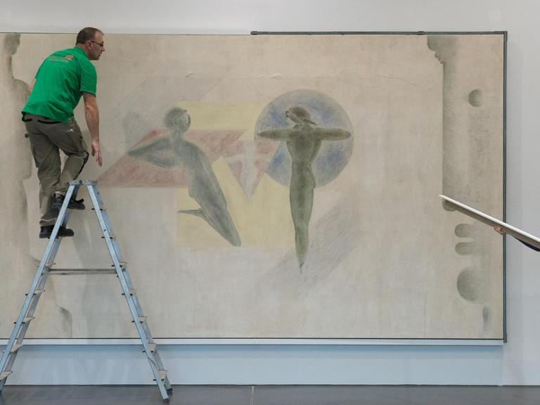 Ein Mann auf einer Leiter und eine Frau mit einem Brett in der Hand stehen vor einem Wandgemälde in einer Galerie.