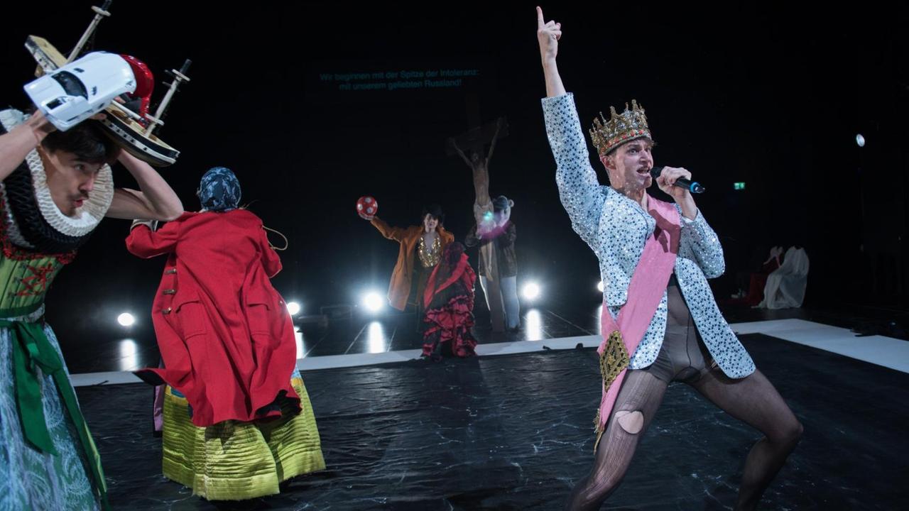 Schauspieler des Schauspiel Stuttgart auf der Bühne in bunten Kostümen. Ein Schauspieler am Bühnenanfang trägt eine Krone und reckt seinen Arm nach oben. Er hält ein Mikrofon und spricht.