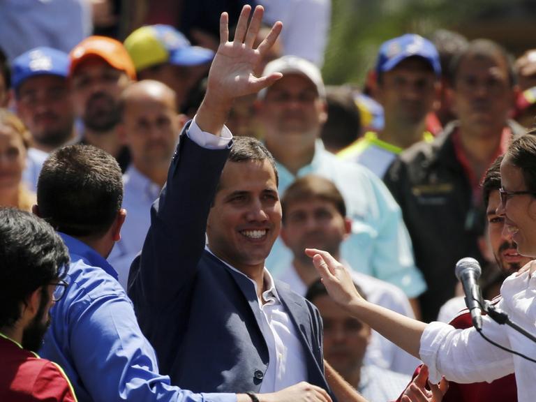 Guaidó steht lächelnd in einer Menschenmenge und winkt; neben ihm steht ein Mikrofon.