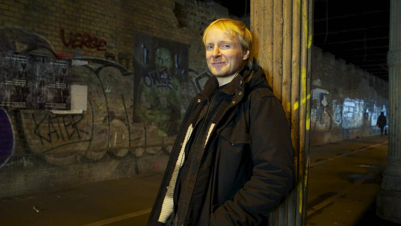 Der Schriftsteller Jochen Schmidt steht unter einer Brücke in der Nacht und blickt verschmitzt in die Kamera.