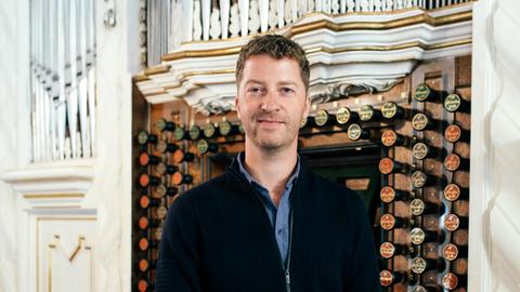 Der Organist steht vor der Orgel (Spieltisch) mit verschiedenen Registerzügen aus Holz. Er hat kurz braune Haare und trägt eine dunkelblaue Strickjacke mit einem jeansfarbenen Hemd.