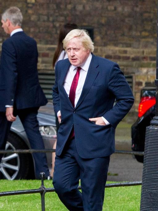 Boris Johnson geht eine Straße entlange, im Hintergrund mehrere Männer und Autos