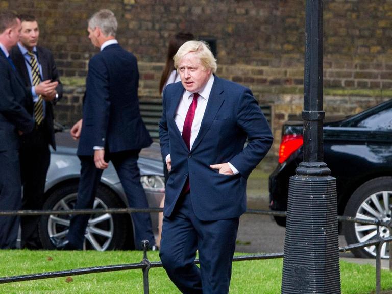 Boris Johnson geht eine Straße entlange, im Hintergrund mehrere Männer und Autos