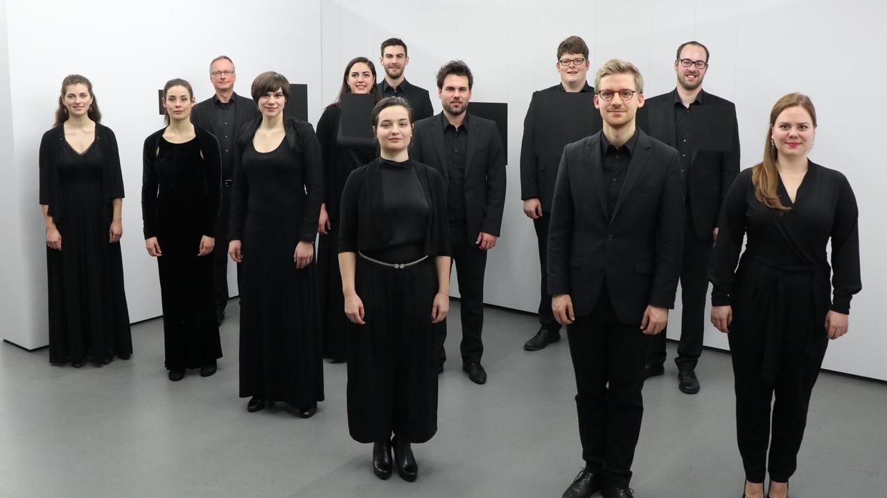 Die Sängerinnen und Sänger von der Jugendchorakademie, alle ganz in schwarz gekleidet, stehen in einem weißen Galerieraum im Raum verteilt und blicken freundlich in die Kamera.