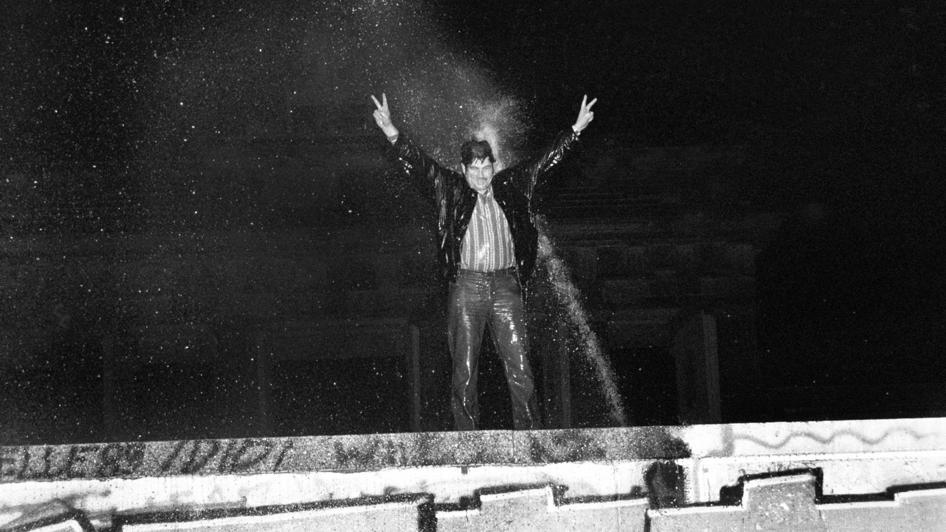 Ein durchnässter Mann steht auf der Berliner Mauer, reckt die Arme in die Höhe und wird dabei vom Strahl eines Wasserwerfers getroffen.