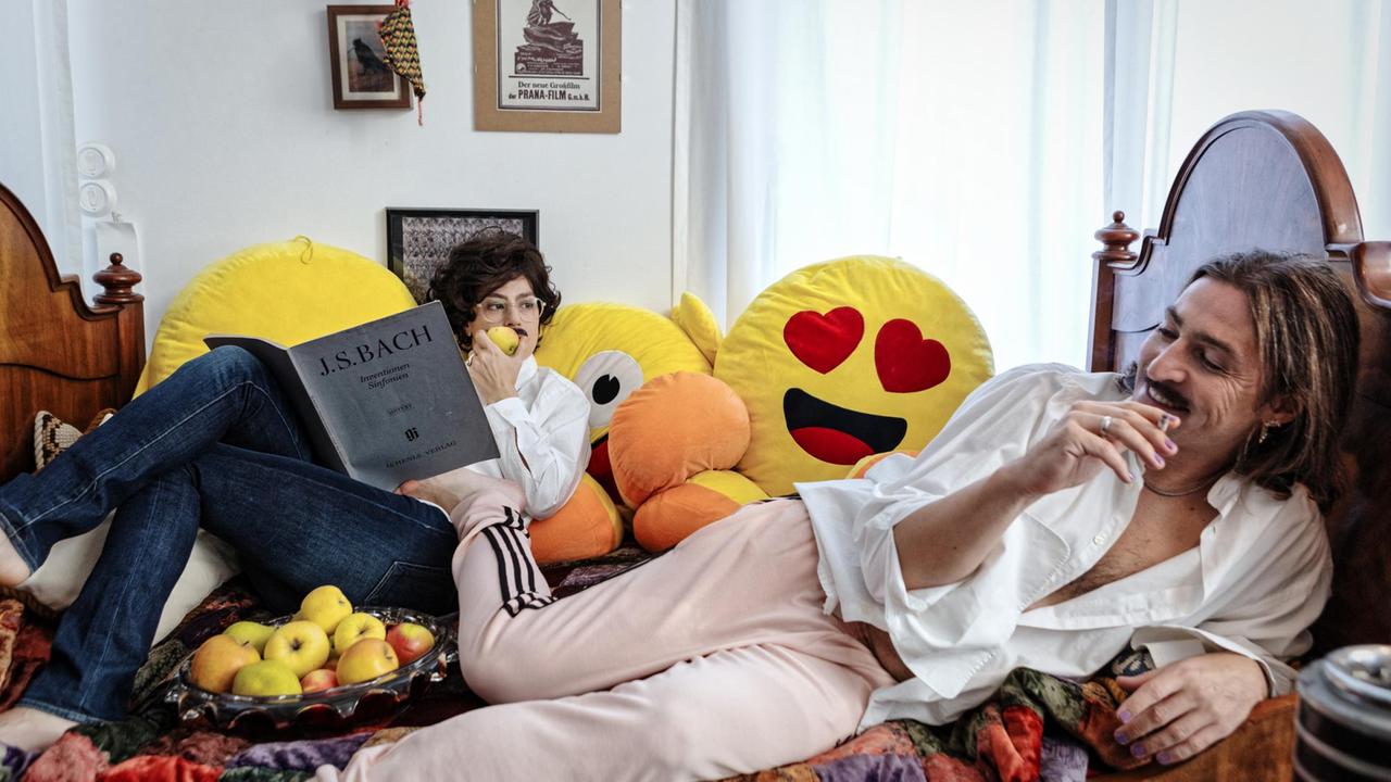 Zwei Menschen sitzen auf einem Bett umgeben von Emoji-Kissen und einem Apfelkorb Zwei Menschen sitzen auf einem Bett umgeben von Emoji-Kissen und einem Apfelkorb
