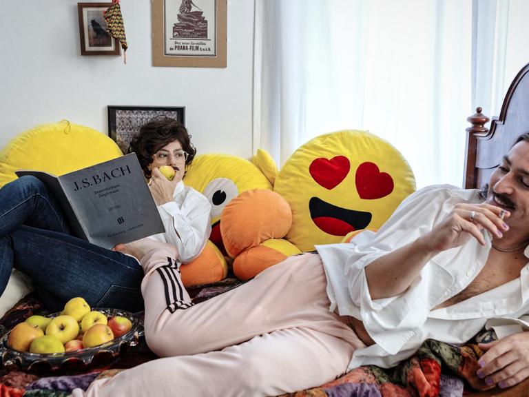 Zwei Menschen sitzen auf einem Bett umgeben von Emoji-Kissen und einem Apfelkorb