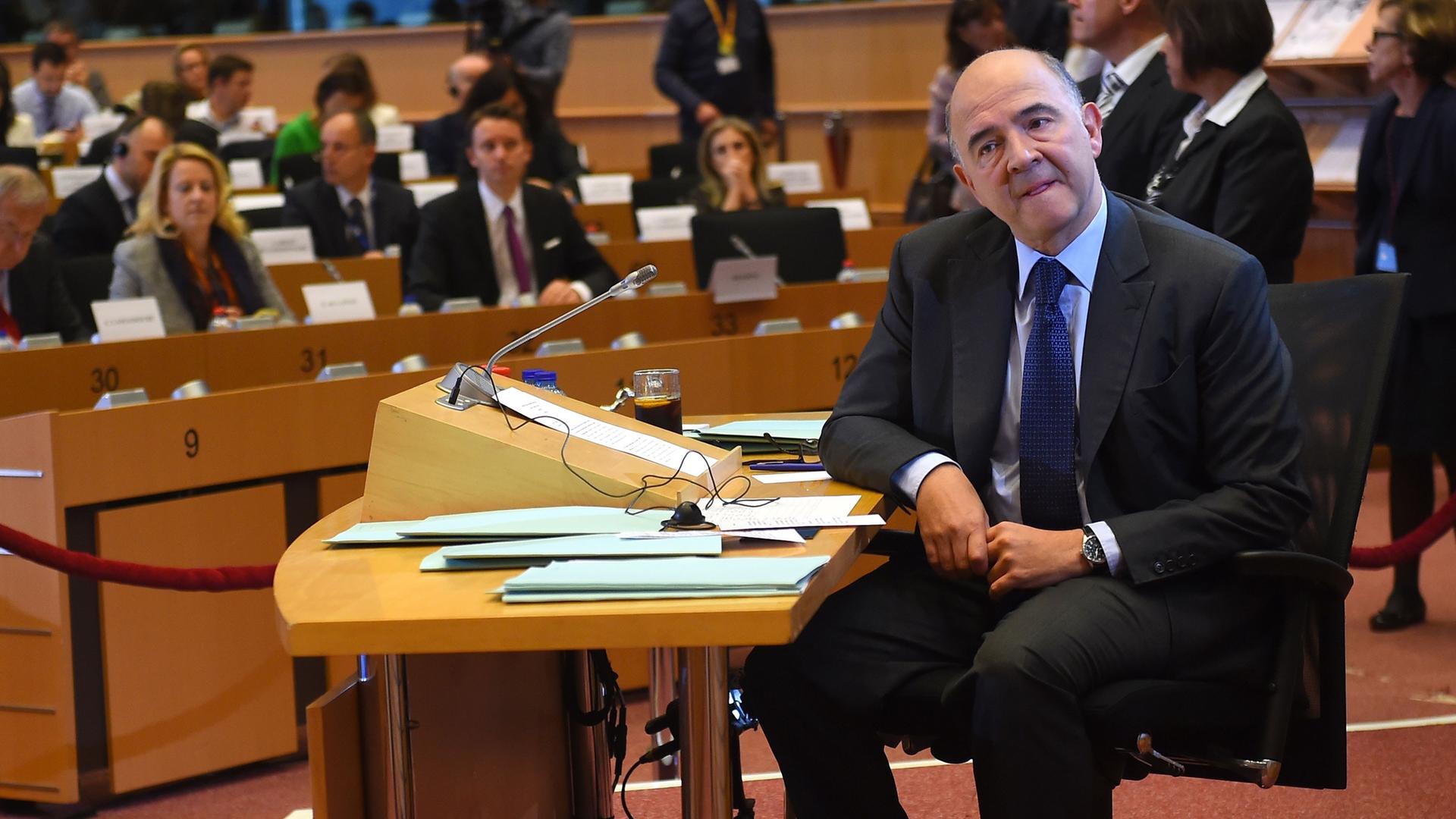 Der Franzose Moscovici sitzt an einem Tisch in einem Ausschuss von dem Europäischen Parlament und schaut in die Kamera. Hinter ihm sieht man Abgeordnete, die ihn befragen wollen. 