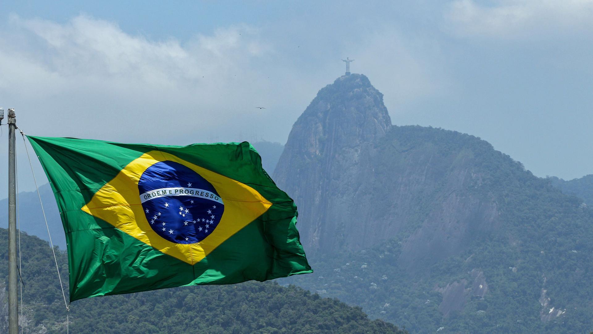 Die brasilianische Flagge. Im Hintergrund: Die monumentale Christusstatue Cristo Redentor auf dem Berg Corcovado in Rio de Janeiro, Brasilien