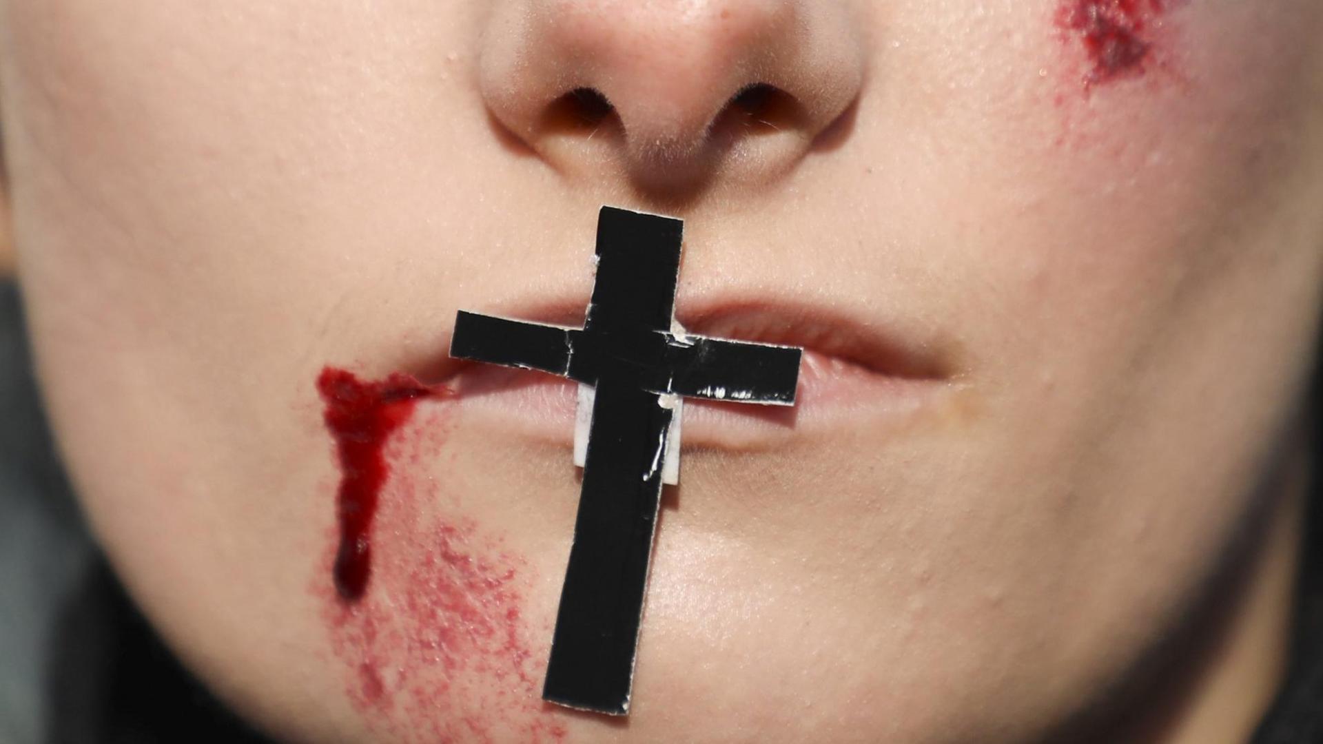 Auf einem Mund klebt ein Kreuz. Im Gesicht sind aufgemalte Wunden zu sehen. Das Bild ist ein Symbolbild für die Folgen von Homophobie in Polen.