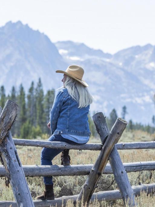Eine Frau mit Cowboyhut sitzt auf einem Holzzaun und beobachtet die Berge in Stanley, Idaho, USA.