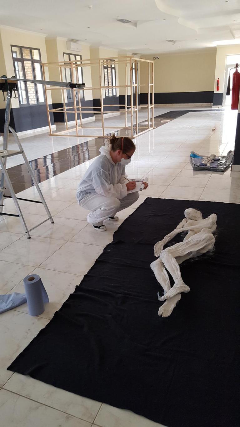 Restauratorin kniet vor dem konservierten Leichnam eines Genozidopfers