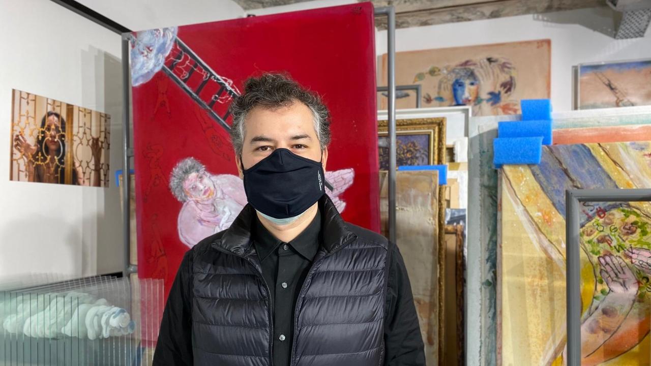 Ein Mann ganz in schwarz mit schwarzem Mundschutz steht in einer Galerie voller bunter Gemälde.