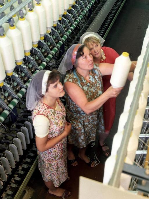 Drei Frauen mit Kittelschürzen und Kopftüchern stehen bei der Arbeit an einer Textilmaschine im VEB Kombinat Baumwolle in der DDR, aufgenommen in den 1980er Jahren