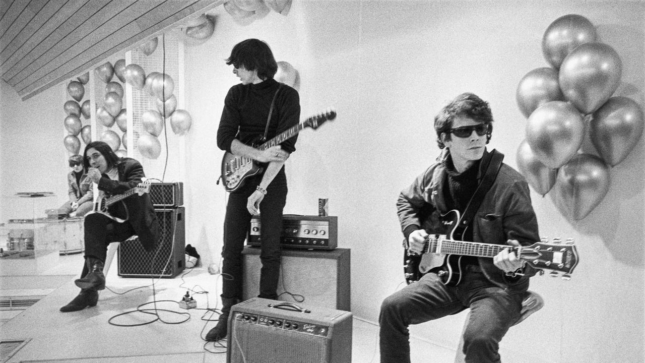 Moe Tucker, John Cale, Sterling Morrison und Lou Reed sitzen beziehungsweise stehen in einem weißen Raum. Sie tragen schwarze Kleidung, Lou Reed rechts eine Sonnenbrille. Sie haben ihre Instrumente und Verstärker bei sich.