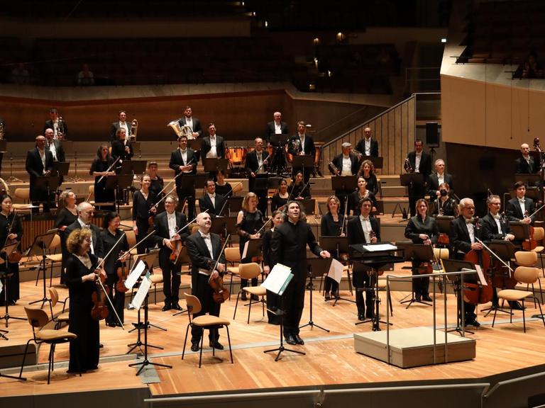 Auf einer Bühne stehen Musikerinnen und Musikers eines Orchesters in schwarzer Konzertkleidung und schauen in Richtung des Publikums, das aber nicht zu sehen ist.