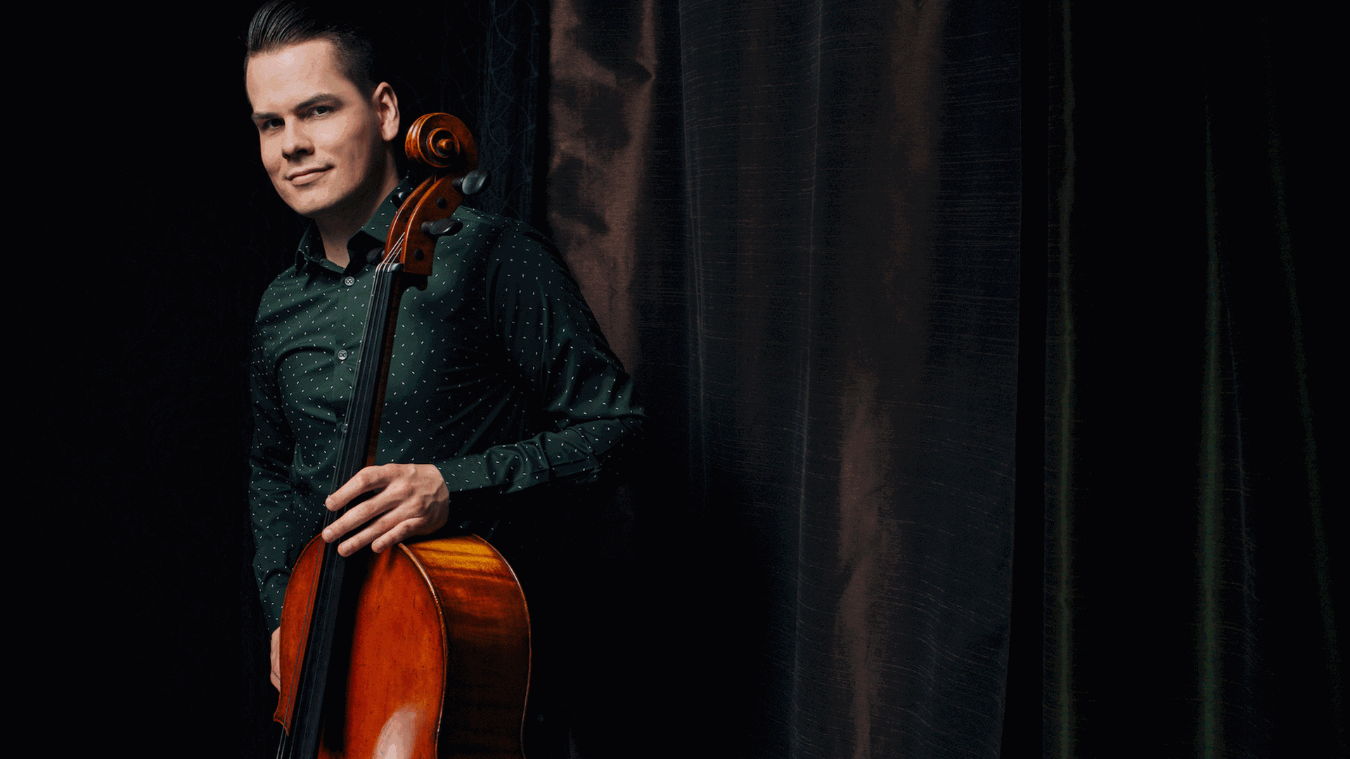 Ein junger Mann mit gegeelten Haaren steht in dunkelgrünem Hemd neben einem dunklen Vorhang mit dem aufgestellten Cello in der Hand, sodass die Schnecke des Instrumentes neben seinem Ohr erscheint.
